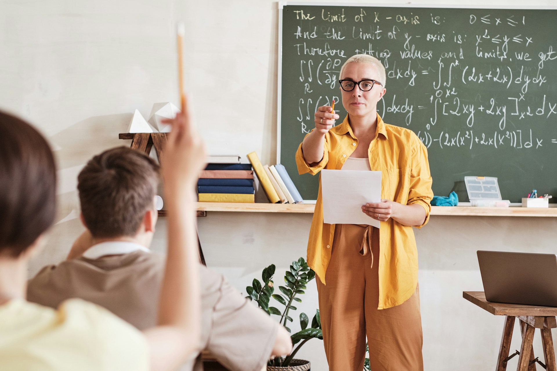 Teacher holding notes in front of a chalkboard with formulas as a learner raises a hand